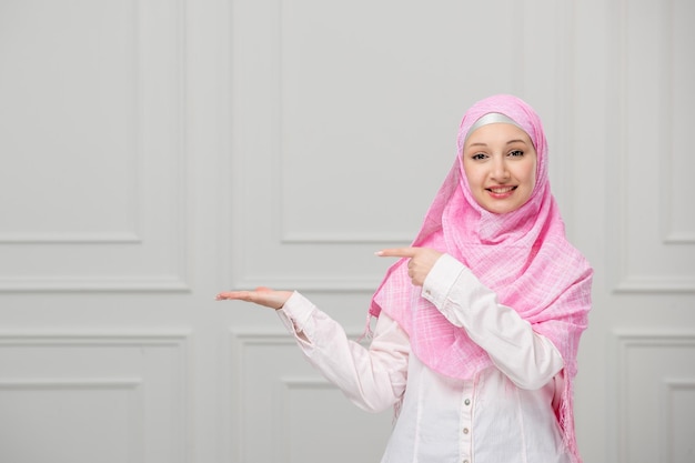 Menina árabe muito bonita jovem muçulmana envolta em lindo hijab rosa apontando para a esquerda
