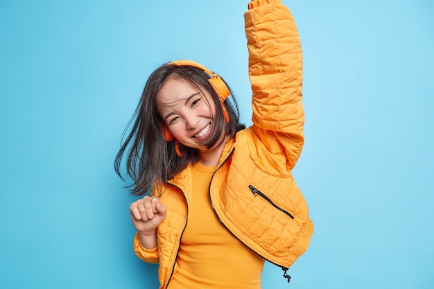 Foto grátis menina asiática alegre se diverte com o cabelo escuro flutuando no ar enquanto pula mantém os braços levantados, usa fones de ouvido sem fio, ouve música, sente-se enérgica isolada sobre a parede azul felicidade do estilo de vida das pessoas