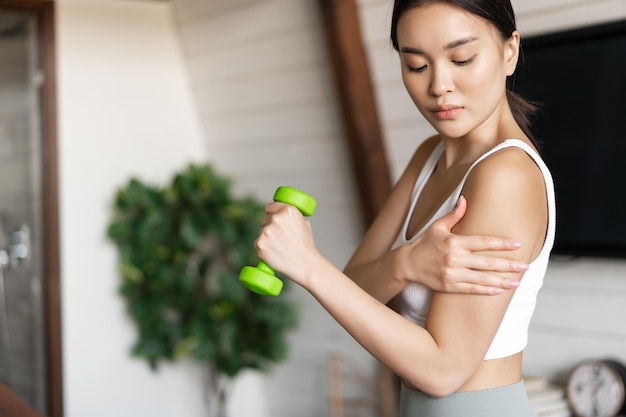 Menina asiática ativa e saudável com corpo em forma, fazendo exercícios de fitness em casa, levantando halteres e ...