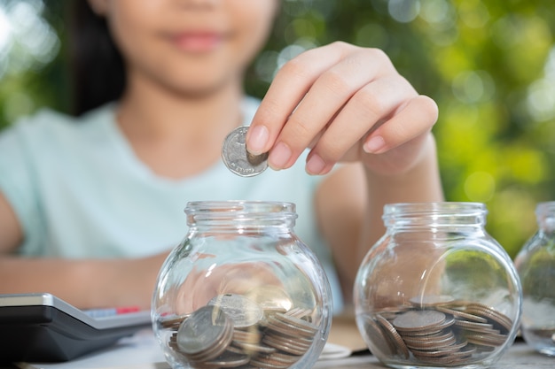 Menina asiática bonitinha brincando com moedas fazendo pilhas de dinheiro, garoto economizando dinheiro no cofrinho, em frasco de vidro. Criança contando suas moedas salvas, crianças aprendendo sobre o conceito de futuro.