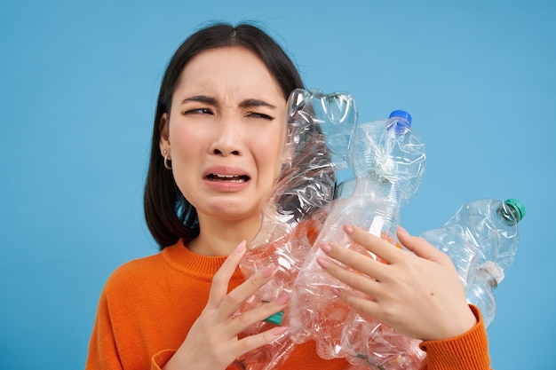 Menina asiática chorando segurando garrafas de plástico sendo chateada reciclando em pé sobre fundo azul