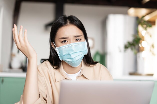 Menina asiática com problemas se sentindo mal usando máscara médica trabalhando em um laptop