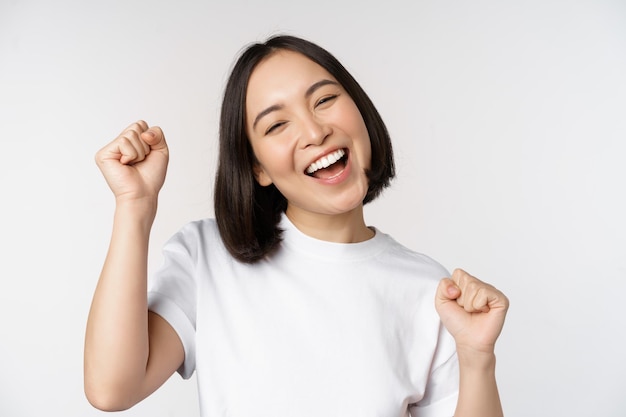 Menina asiática dançando comemorando se sentindo feliz e otimista sorrindo amplamente em pé sobre o fundo branco do estúdio
