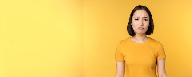 Menina asiática desapontada, amuada, parecendo chateada, sente-se sem ar em camiseta amarela sobre fundo branco