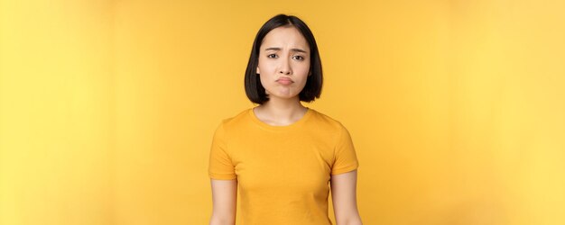 Menina asiática desapontada, amuada, parecendo chateada, sente-se sem ar em camiseta amarela sobre fundo branco