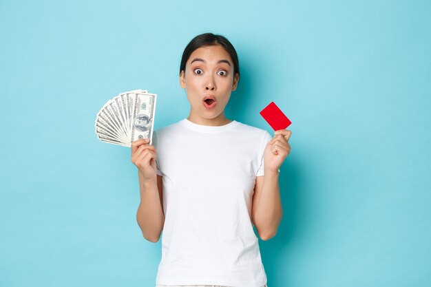 Menina asiática divertida em uma camiseta casual branca arfando, descobriu preços incríveis, ofertas de desconto na loja, segurando cartão de crédito e dinheiro, parede azul claro