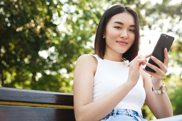 Menina asiática elegante sentada no parque e usando o smartphone, sorrindo para a câmera