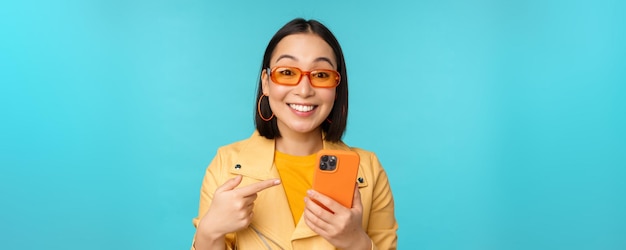 Menina asiática entusiasmada em óculos de sol elegantes apontando o dedo para o smartphone mostrando o celular