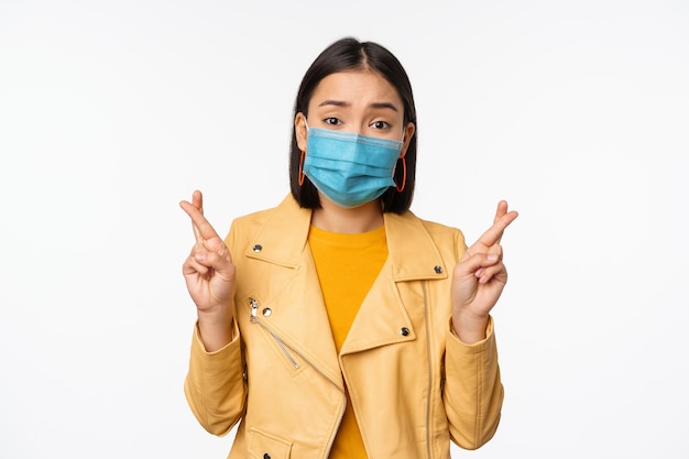 Menina asiática esperançosa em máscara facial médica cruzar os dedos fazendo um desejo esperando orar por algo em pé com antecipação sobre o fundo branco do estúdio