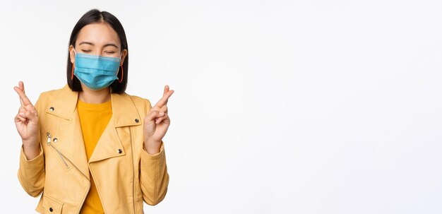 Menina asiática esperançosa em máscara facial médica cruzar os dedos fazendo um desejo esperando orar por algo em pé com antecipação sobre o fundo branco do estúdio