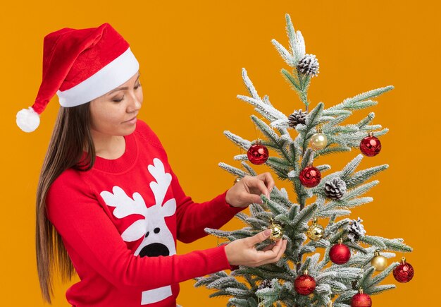 Menina asiática feliz com chapéu de Natal e suéter decorando a árvore de Natal isolada na parede laranja