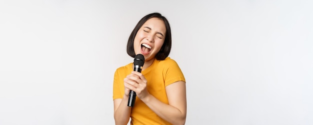 Menina asiática feliz dançando e cantando karaokê segurando o microfone na mão se divertindo em pé sobre fundo branco