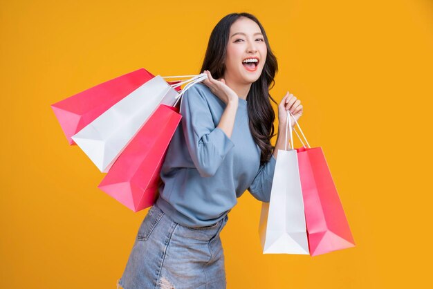 Menina asiática feliz mulher segura pacotes de compras coloridos em pé no estúdio de fundo amarelo Feche o retrato jovem linda garota atraente sorrindo olhando para a câmera com sacos