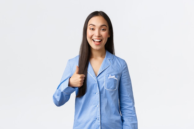 Menina asiática feliz otimista de pijama azul sorrindo satisfeita e mostrando o polegar para cima em aprovação ou tipo, recomendar produto, ótima qualidade, mostrando bem feito ou bom trabalho, fundo branco.