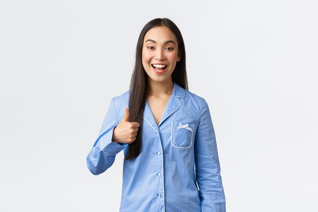 Menina asiática feliz otimista de pijama azul sorrindo satisfeita e mostrando o polegar para cima em aprovação ou tipo, recomendar produto, ótima qualidade, mostrando bem feito ou bom trabalho, fundo branco.