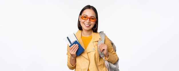 Menina asiática feliz saindo de férias segurando mochila de passaporte e passagens aéreas no ombro Jovem turista viajando para o exterior em pé sobre fundo branco