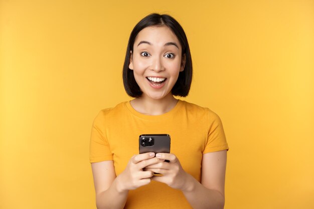 Menina asiática feliz sorrindo em pé com o celular preto em pé contra o fundo amarelo