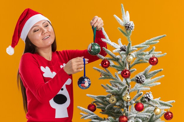 Menina asiática feliz usando um chapéu de Natal e um suéter decorando uma árvore de Natal isolada em um fundo laranja