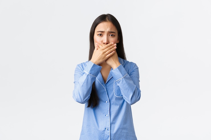 Foto grátis menina asiática fofa preocupada de pijama azul franzindo a testa e parece preocupada enquanto cala a boca com as mãos, tampa os lábios, reagindo a notícias terríveis, em pé, fundo branco chateado.