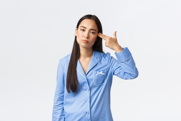 Menina asiática irritada e incomodada de pijama azul parecendo relutante, atirando em si mesma com o gesto de uma arma como se sentindo farta, cansada de ouvir ou ver algo chato ou idiota, fundo branco.