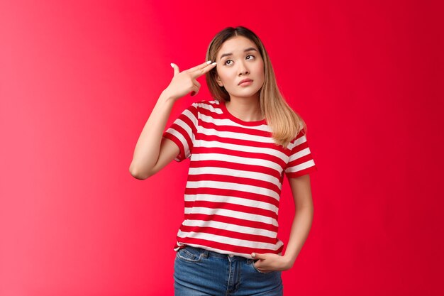 Menina asiática loira chateada com camiseta listrada jeans morrendo de tédio, suspirando, olhar para cima, segurar o dedo, arma pisto ...