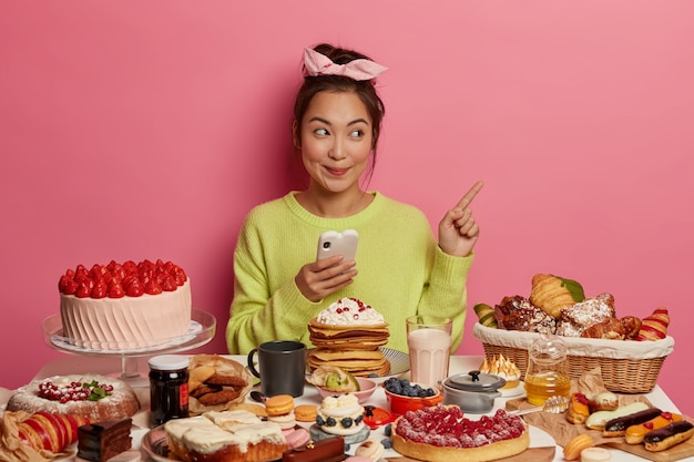 Foto grátis menina asiática morena alegre alegre rodeada de biscoitos, biscoitos e bolos, gosta de comida doce durante o tempo festivo, gosta de guloseimas de férias, aponta para o espaço em branco, usa o telefone celular.