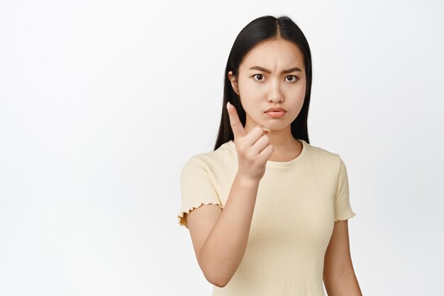 Menina asiática morena irritada balançando o dedo para desaprovar smth repreendendo o mau comportamento de pé na camiseta sobre fundo branco