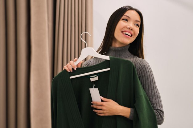 Menina asiática muito alegre tentando felizmente um casaco de lã na loja de moda moderna