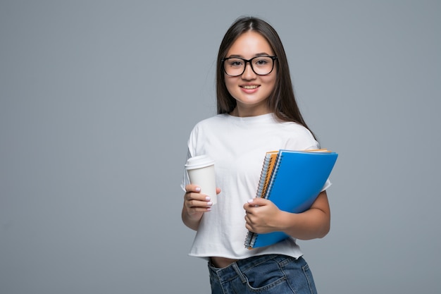 Menina asiática nova com caderno e café para ir nas mãos que estão isoladas contra o fundo cinzento