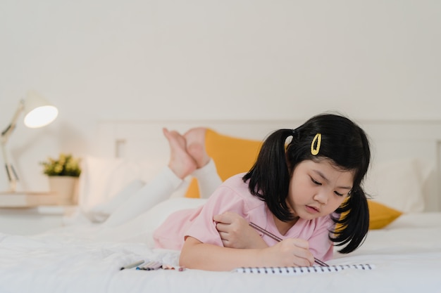 Menina asiática nova que tira em casa. a criança japonesa da criança da mulher de ásia relaxa desenhos animados felizes da tração do divertimento do resto no caderno antes de dormir deitado na cama, sinta o conforto e a calma no quarto no conceito da noite.