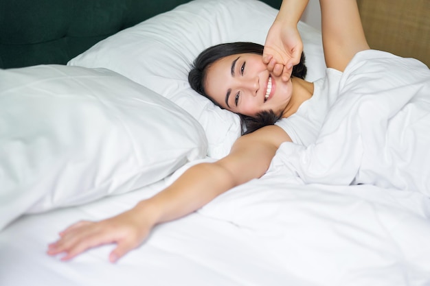 Menina asiática romântica acordando em um quarto aconchegante, deitada na cama com lençóis brancos esticando a mão