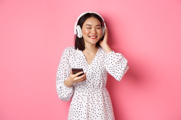 Menina asiática romântica ouvindo música em fones de ouvido, sorrindo com os olhos fechados, segurando um telefone celular, em pé sobre um fundo rosa