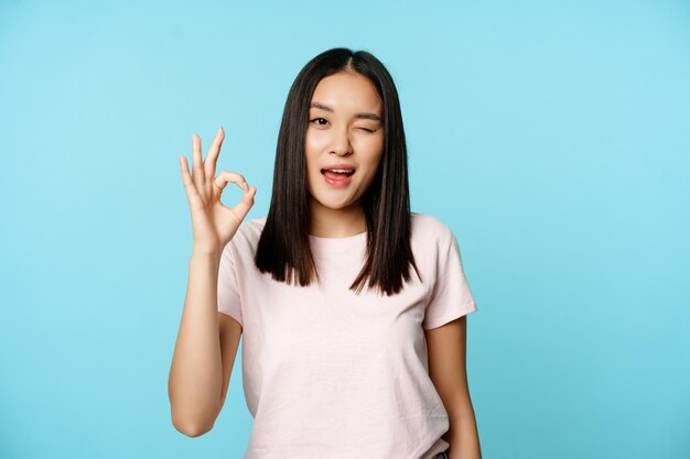 Menina asiática satisfeita pisca o olho e mostra sinal de ok dar aprovação, recomenda estandes em camiseta sobre bac azul ...
