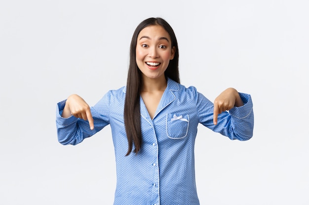 Menina asiática sorridente alegre reage a notícias maravilhosas, apontando os dedos para baixo como em pé de pijama azul, mostrando coisa legal para namoradas na festa do pijama, fazendo o anúncio, fundo branco.