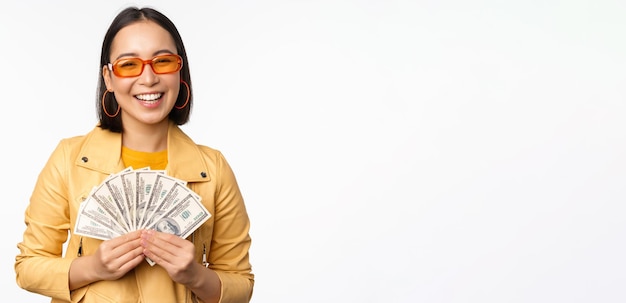 Menina asiática sorridente elegante segurando dinheiro mostrando dólares e comemorando em pé sobre fundo branco Copie o espaço