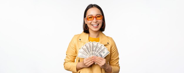 Menina asiática sorridente elegante segurando dinheiro mostrando dólares e comemorando em pé sobre fundo branco