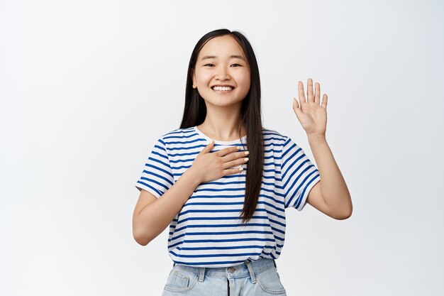 Menina asiática sorridente feliz levantando uma mão, colocando a palma no coração, xingando, fazendo uma promessa, apresentando-se, pisando em branco