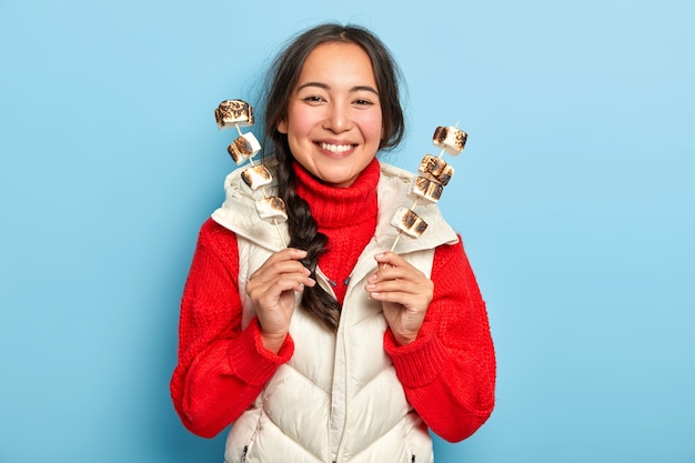 Menina asiática sorridente feliz segurando palitos com deliciosos marshmallows assados aromáticos, gosta de fazer um piquenique no campo e usa roupas quentes
