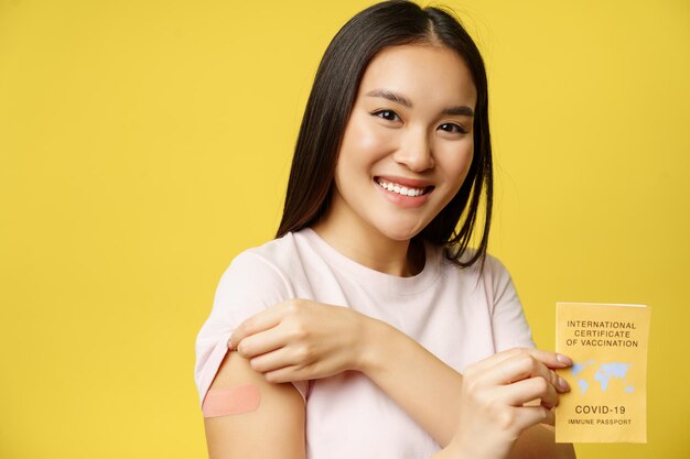 Menina asiática sorridente mostrando certificado internacional de vacinação covid 19, demonstrando braço com adesivo após a vacina de coronavírus, fundo amarelo