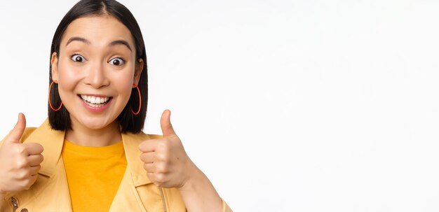 Menina asiática sorridente mostrando os polegares para cima parecendo satisfeita aprovar algo sobre fundo branco