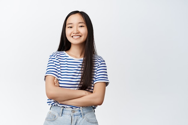 Menina asiática sorridente parecendo feliz, braços cruzados no peito, em pé com uma camiseta listrada em branco