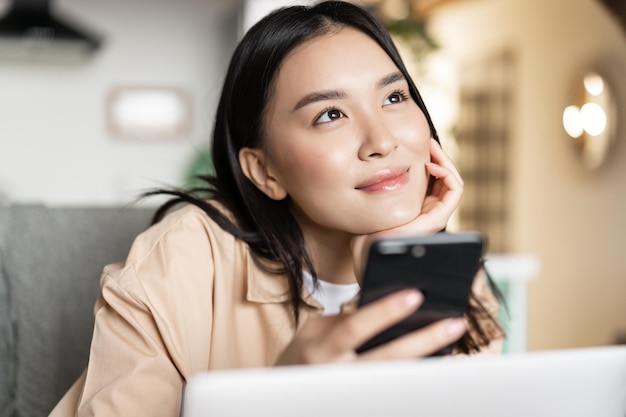 Menina asiática sorridente segurando o telefone e pensando, parecendo um sonho, sentada com o laptop em casa