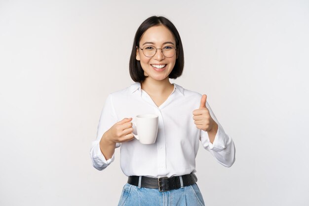 Menina asiática sorridente segurando xícara branca de caneca e polegares para cima recomendando beber café ou chá em pé sobre fundo branco