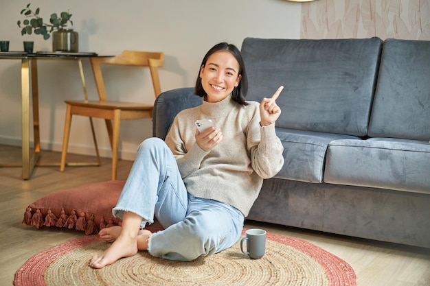 Foto grátis menina asiática sorridente sentada no chão em uma elegante sala de estar apontando o dedo para exibição de anúncio