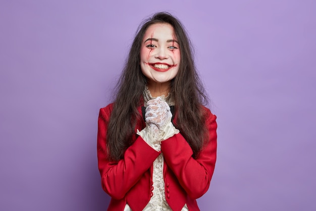 Menina assustadora de halloween com sorrisos assustadores de maquiagem antecipa alegremente para a festa de máscaras mantém as mãos juntas isoladas na parede roxa. arte de rosto sangrento