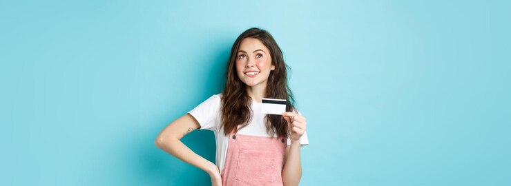 Foto grátis menina atenciosa pensando em fazer compras segurando cartão de crédito de plástico e olhando para cima com rosto pensativo fazendo escolha o que comprar de pé sobre fundo azul
