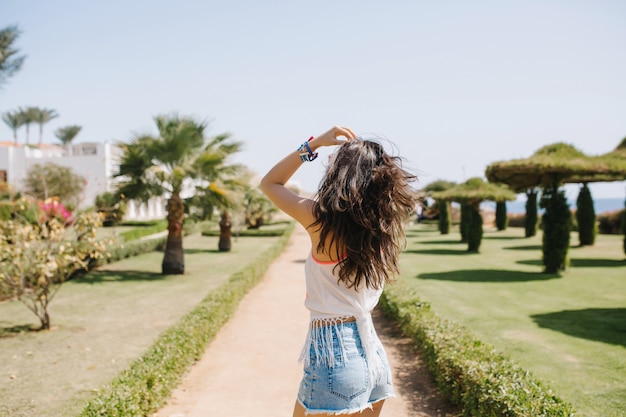 Menina bem torneada de cabelos compridos em camisa branca, caminhando no beco sob o céu azul na manhã ensolarada. Retrato de engraçado jovem elegante dançando no parque do resort nas férias de verão.