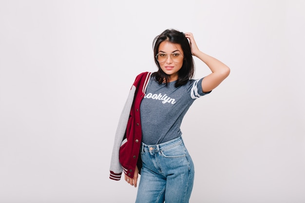 Menina bem torneada em jeans e óculos elegantes, posando sensualmente. Retrato interior da adorável modelo feminino com cabelo escuro curto isolado.