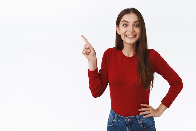 Menina bonita alegre e divertida mostrando a você uma promoção incrível, apontando no canto superior esquerdo, segurando a mão no quadril e sorrindo amplamente com espanto e curiosidade, em pé com um fundo branco entusiasmado