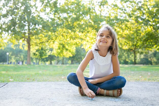 Menina bonita alegre sentada e desenhando com pedaços coloridos de giz, olhando para longe e sorrindo. Vista frontal. Conceito de infância e criatividade